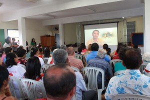 Participantes das festividades de Nossa Senhora da Conceição assistem às apresentações das videocabines, algumas delas gravadas no distrito, durante a 8ª exibição da Mostra de Videocabines – “Festas Populares II”, no Salão Paroquial da Igreja de Nossa Senhora da Conceição, no distrito de Cachoeira do Brumado, em Mariana. Foto: Wesley Maurício