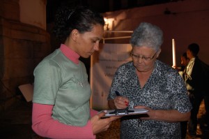 A depoente Maria Aparecida Santos, assina o Termo de Autorização para uso de imagem, ao lado de Cristiane Evangelista, no adro da Igreja Nossa Senhora do Rosário dos Pretos, no bairro Rosário, em Ouro Preto. Foto: Marianne Fonseca 